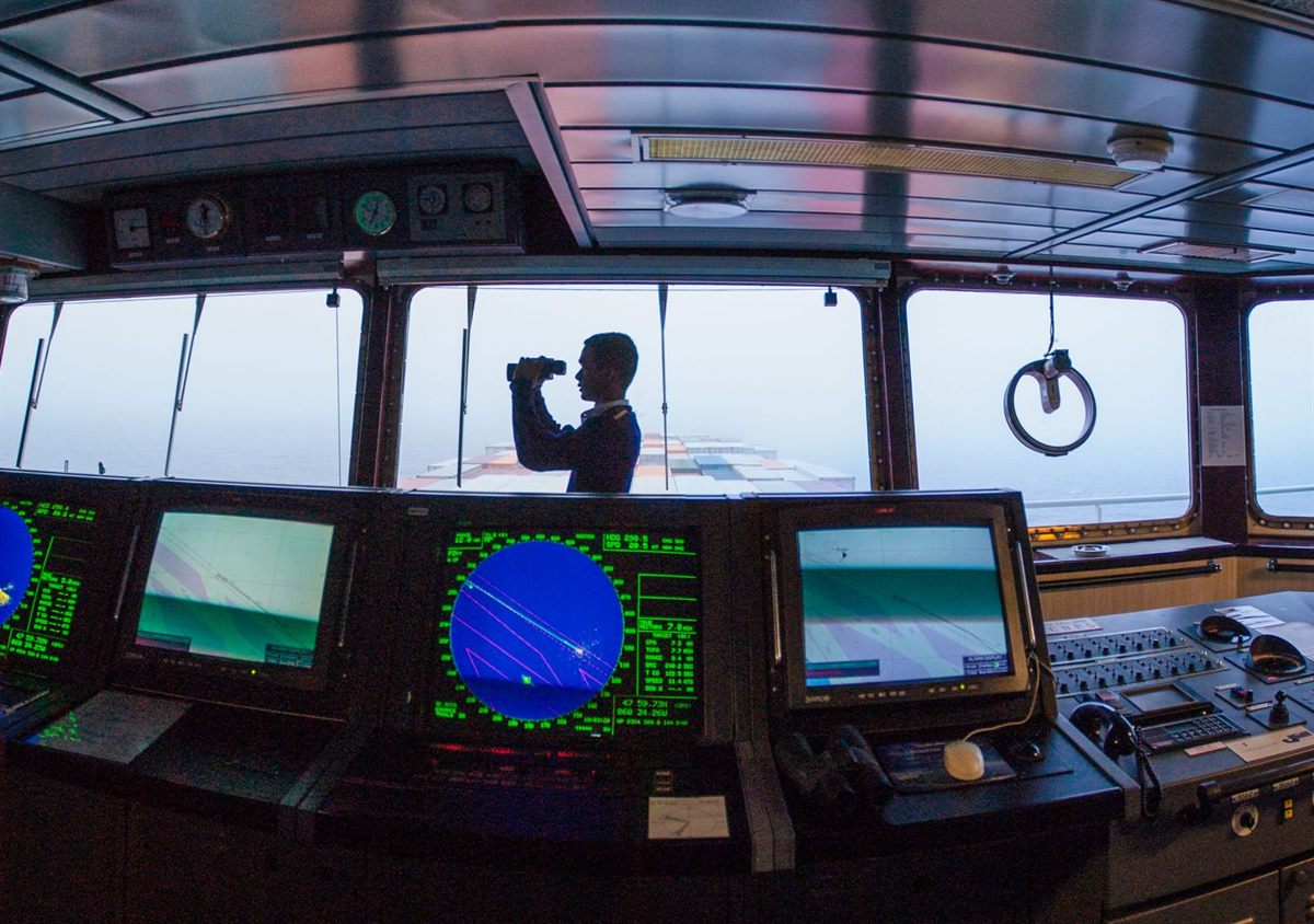 An officer with binoculars on a ship bridge surrounded by radar and navigation screens